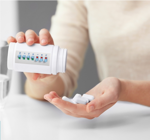 The image depicts a person's hands in the process of taking medication, with a focus on a white pill bottle equipped with a medication reminder system.