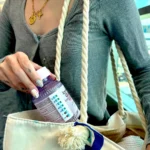 Woman taking a supplement bottle with a Take-n-Slide medication tracker out of her travel bag, showing colorful nails and a casual outfit